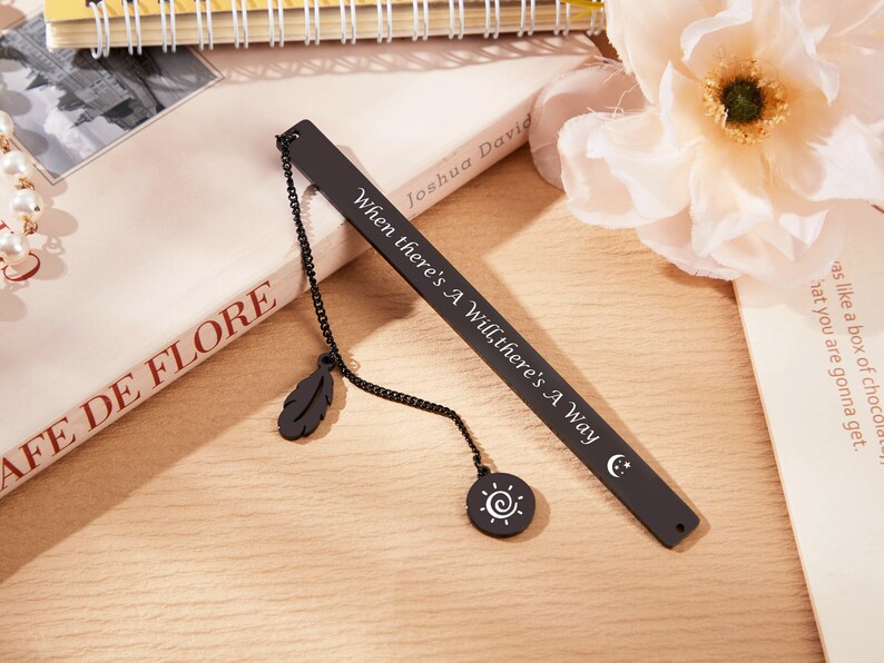 May include: Black metal bookmark with a chain and two charms, a feather and a sun. The bookmark is engraved with the quote "When there's a will, there's a way."