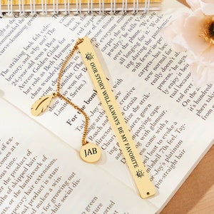 May include: A gold bookmark with the text "Our story will always be my favorite" and a sun symbol. The bookmark is on a book with a gold chain and a feather charm.