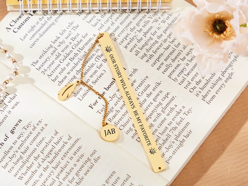 May include: A gold bookmark with the text "Our story will always be my favorite" and a sun symbol. The bookmark is on a book with a gold chain and a feather charm.