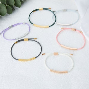 May include: A collection of six bracelets in various colors, including purple, green, blue, pink, gray, and white. Each bracelet features a beaded design with a metallic bar and clasp. The bracelets are displayed on a white surface.