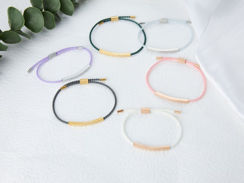 May include: A collection of six bracelets in various colors, including purple, green, blue, pink, gray, and white. Each bracelet features a beaded design with a metallic bar and clasp. The bracelets are displayed on a white surface.