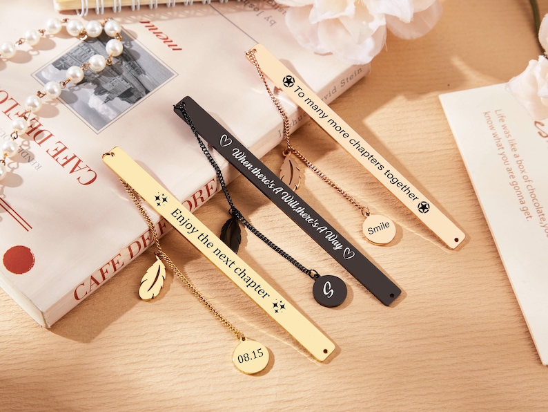 May include: Set of five gold and black metal bookmarks with different inspirational quotes and charms. The bookmarks feature a feather charm, a star charm, a heart charm, and a circle charm. The quotes include "To many more chapters together", "When there's a will, there's a way", "Enjoy the next chapter", and "Smile".