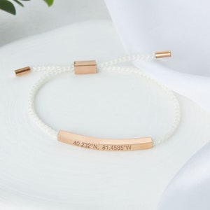 May include: White cord bracelet with rose gold accents. The bracelet features a rose gold bar engraved with coordinates: 40.232&deg;N, 81.4585&deg;W. The bracelet has a sliding knot closure for adjustable sizing.