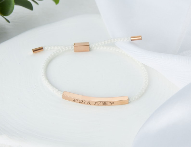 May include: White cord bracelet with rose gold accents. The bracelet features a rose gold bar engraved with coordinates: 40.232&deg;N, 81.4585&deg;W. The bracelet has a sliding knot closure for adjustable sizing.