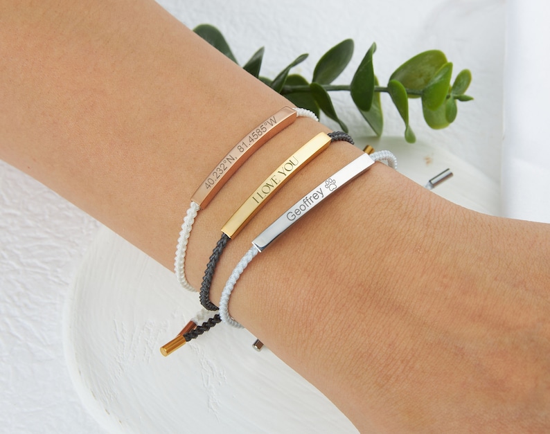 May include: Three personalized bar bracelets in rose gold, gold, and silver. The rose gold bracelet has coordinates, the gold one says "I LOVE YOU", and the silver one has a paw print and the name "Geoffrey". Each bracelet has a braided cord.