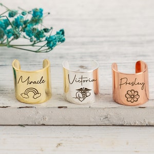 Op de afbeelding: Drie gepersonaliseerde manchet armbanden met verschillende ontwerpen. De eerste armband is goud en gegraveerd met het woord "Miracle" en een regenboog. De tweede armband is zilver en gegraveerd met het woord "Victoria" en een medisch symbool. De derde armband is koper en gegraveerd met het woord "Presley" en een bloem.