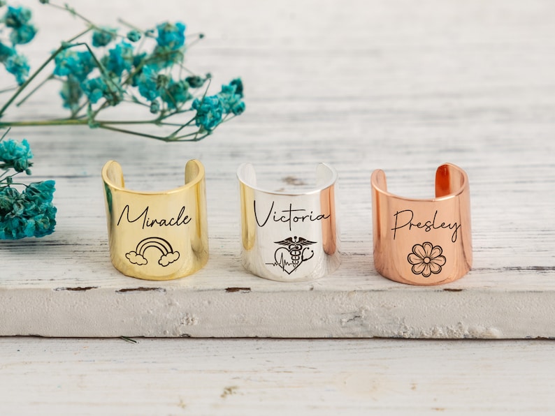 Op de afbeelding: Drie gepersonaliseerde manchet armbanden met verschillende ontwerpen. De eerste armband is goud en gegraveerd met het woord "Miracle" en een regenboog. De tweede armband is zilver en gegraveerd met het woord "Victoria" en een medisch symbool. De derde armband is koper en gegraveerd met het woord "Presley" en een bloem.
