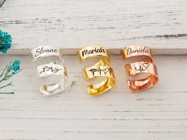 Op de afbeelding: Drie gepersonaliseerde, gegraveerde ringen van zilver, goud en ros&eacute;goud met de namen Sloane, Mariah en Daniela. Elke ring heeft een ander ontwerp, waaronder een tak, een stethoscoop en een hartslag, en een hart met een hartslag.