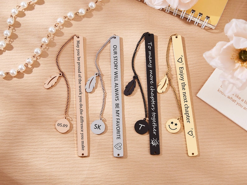 May include: Four gold, silver, and black metal bookmarks with different engraved messages and charms. The bookmarks have a feather charm and a small round charm with different engravings. The messages on the bookmarks are "May you be proud of the work you do, the difference you make", "OUR STORY WILL ALWAYS BE MY FAVORITE", "To many more chapters together", and "Enjoy the next chapter".