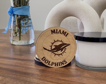 May include: A wooden coaster with the text "Miami Dolphins" and a stylized dolphin logo. The coaster is brown and has a natural wood grain.
