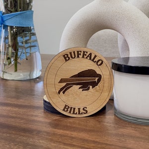 Könnte beinhalten: Runde Holzuntersetzer mit dem Logo der Buffalo Bills und der Aufschrift "BUFFALO BILLS". Die Untersetzer haben eine hellbraune Oberfläche. Ein Stapel Untersetzer ist sichtbar.