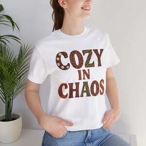 Puede incluir: Camiseta blanca con el texto "COZY IN CHAOS" en marrón, beige y verde oliva. La palabra "COZY" tiene un diseño de estampado de leopardo y lunares. Una prenda casual para el día a día.