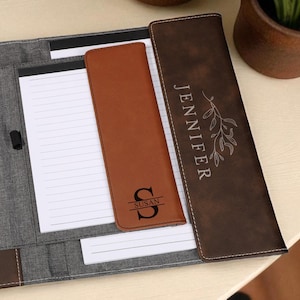 May include: Open leather portfolio with a gray fabric interior. Features a brown leather pocket and pen holder. Includes a notepad and a brown leather pen holder with the letter "S". The portfolio is personalized with the name "Jennifer".