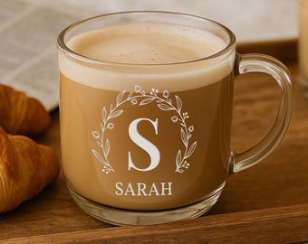Op de afbeelding: Een heldere glazen mok gevuld met koffie, met een witte bloemenkrans met de letter "S" en de naam "Sarah". De mok staat op een houten dienblad met twee croissants en een brandende kaars.