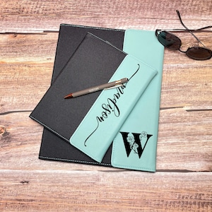 Op de afbeelding: Een teal en zwart lederen notitieboek met een gepersonaliseerde inscriptie en een bloemige initial "W" op de voorzijde. Een pen ligt op het notitieboek.