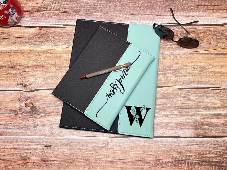 Op de afbeelding: Een teal en zwart lederen notitieboek met een gepersonaliseerde inscriptie en een bloemige initial "W" op de voorzijde. Een pen ligt op het notitieboek.