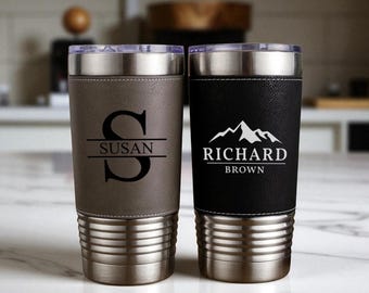 May include: Two stainless steel insulated tumblers with lids. One is gray with the name "Susan" and a large initial. The other is black with the name "Richard Brown" and a mountain graphic. Both have a silver base.