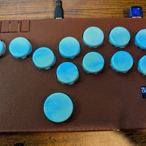 May include: A brown arcade controller with twelve light blue buttons and a small screen displaying a series of blue circles. The controller has a rectangular shape and a USB cable.