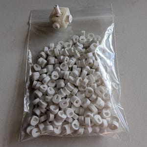 May include: A clear plastic bag filled with small, white, cylindrical objects and a larger, off-white, geometric shape. The cylindrical objects have a hole in the center. The geometric shape has multiple protruding cylinders.