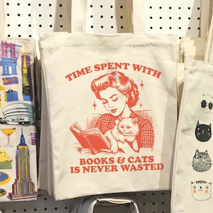 May include: A white canvas tote bag with a red graphic of a woman reading a book with a cat in her lap. The text on the bag reads "Time Spent With Books & Cats Is Never Wasted."