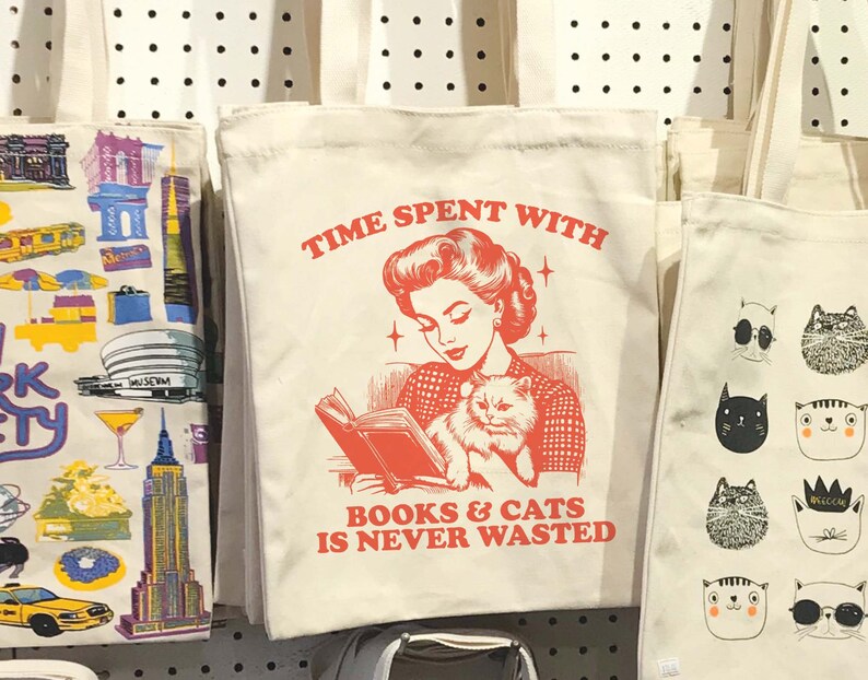May include: A white canvas tote bag with a red graphic of a woman reading a book with a cat in her lap. The text on the bag reads "Time Spent With Books & Cats Is Never Wasted."