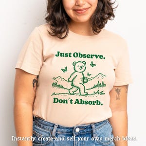 May include: A light beige t-shirt with a green graphic of a bear walking in a forest with butterflies. The text "Just Observe. Don't Absorb." is printed on the shirt.