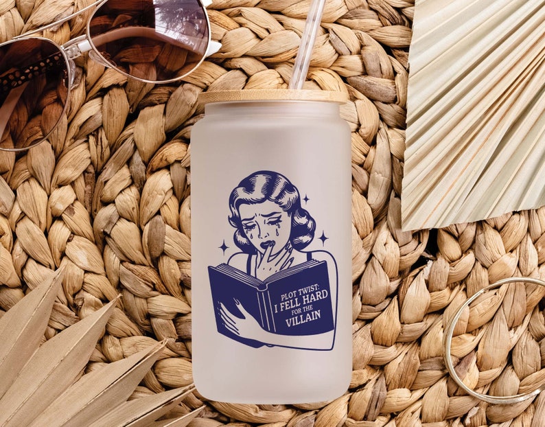 May include: A white reusable water bottle with a blue and white illustration of a woman reading a book. The text on the book reads "Plot Twist: I Fell Hard for the Villain".