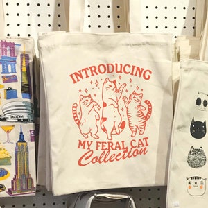 Puede incluir: Tres bolsas de tela blancas con diferentes dise&ntilde;os. La bolsa del centro tiene texto rojo que dice "Introducing My Feral Cat Collection" con tres gatos de dibujos animados bailando. La bolsa de la izquierda tiene un dise&ntilde;o colorido de la ciudad de Nueva York. La bolsa de la derecha tiene un dise&ntilde;o de gato blanco y negro.