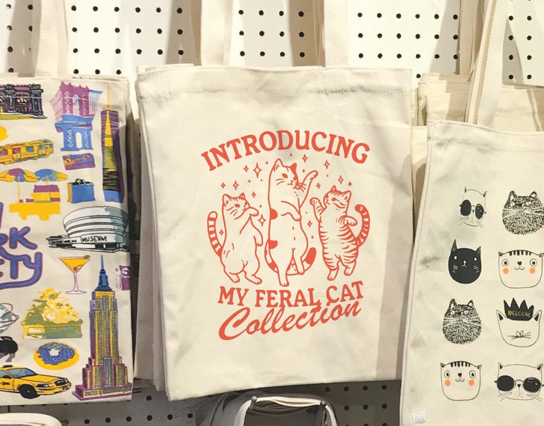 Puede incluir: Tres bolsas de tela blancas con diferentes dise&ntilde;os. La bolsa del centro tiene texto rojo que dice "Introducing My Feral Cat Collection" con tres gatos de dibujos animados bailando. La bolsa de la izquierda tiene un dise&ntilde;o colorido de la ciudad de Nueva York. La bolsa de la derecha tiene un dise&ntilde;o de gato blanco y negro.