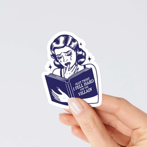 May include: A blue and white sticker featuring a cartoon woman reading a book. The book reads "Plot Twist: I Fell Hard For The Villain".