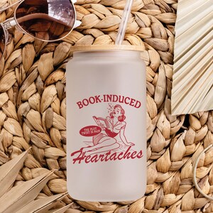 May include: A white frosted glass tumbler with a bamboo lid and a red graphic that says "Book-Induced Heartaches" with a cartoon illustration of a woman reading a book. The text below the illustration says "The plot twist is pain."