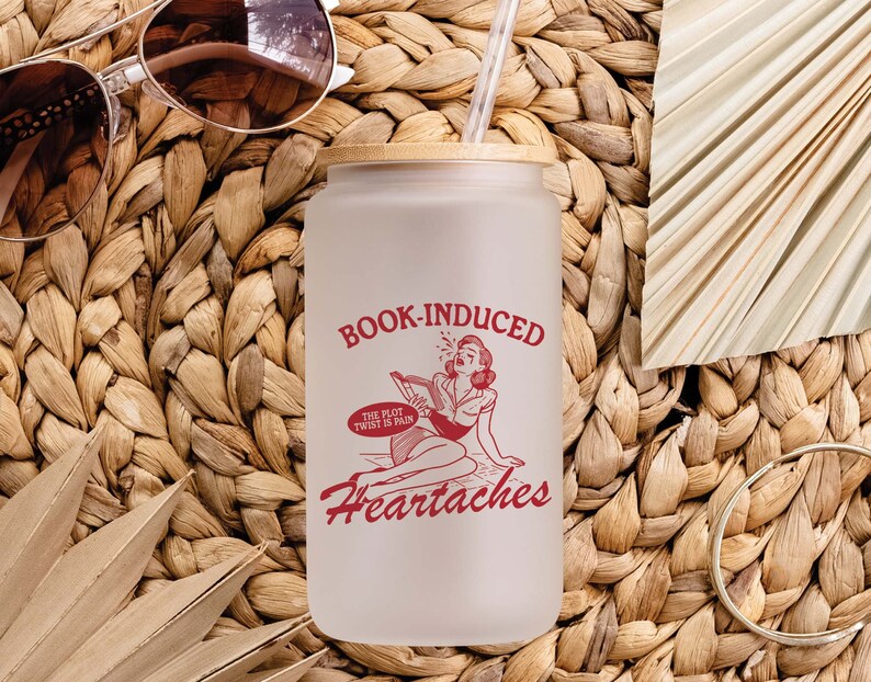 May include: A white frosted glass tumbler with a bamboo lid and a red graphic that says "Book-Induced Heartaches" with a cartoon illustration of a woman reading a book. The text below the illustration says "The plot twist is pain."