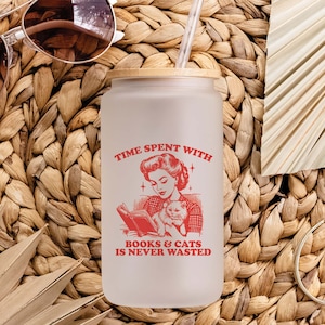 May include: A frosted glass water bottle with a bamboo lid and a red graphic that says "Time spent with books & cats is never wasted". The graphic features a woman reading a book with a cat in her lap.