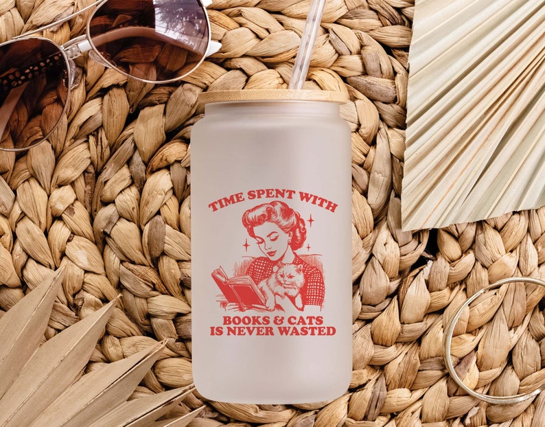 May include: A frosted glass water bottle with a bamboo lid and a red graphic that says "Time spent with books & cats is never wasted". The graphic features a woman reading a book with a cat in her lap.