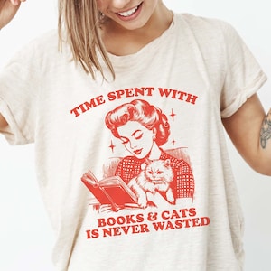 May include: A white t-shirt with a red graphic of a woman reading a book with a cat in her lap. The text on the shirt reads "Time spent with books & cats is never wasted."