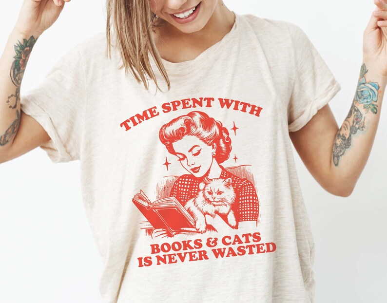 May include: A white t-shirt with a red graphic of a woman reading a book with a cat in her lap. The text on the shirt reads "Time spent with books & cats is never wasted."