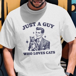 May include: White t-shirt featuring a retro-style illustration of a man holding a cat. The text "Just a guy who loves cats" is printed on the shirt.