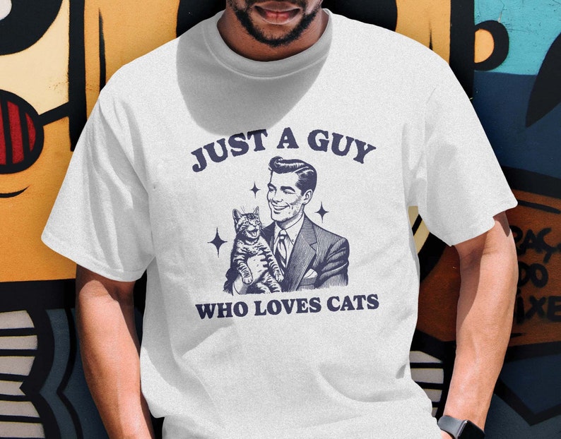 May include: White t-shirt featuring a retro-style illustration of a man holding a cat. The text "Just a guy who loves cats" is printed on the shirt.