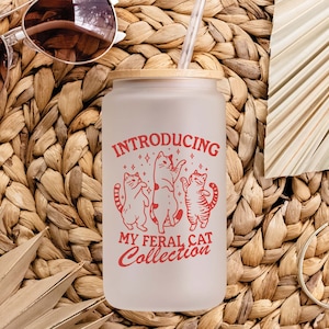 Puede incluir: Un vaso de vidrio esmerilado con tapa de bamb&uacute; y un gr&aacute;fico rojo que dice "Introducing My Feral Cat Collection". El gr&aacute;fico presenta tres gatos estilizados bailando con estrellas a su alrededor.