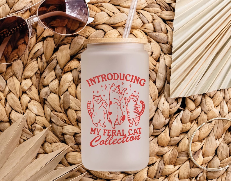 Puede incluir: Un vaso de vidrio esmerilado con tapa de bamb&uacute; y un gr&aacute;fico rojo que dice "Introducing My Feral Cat Collection". El gr&aacute;fico presenta tres gatos estilizados bailando con estrellas a su alrededor.