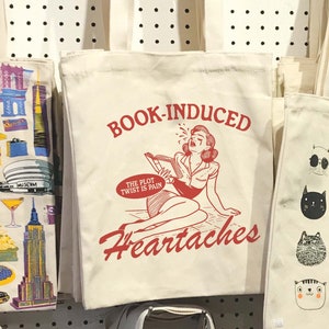 May include: A white canvas tote bag with a red graphic of a woman reading a book and the text "BOOK-INDUCED HEARTACHES" and "THE PLOT TWIST IS PAIN".