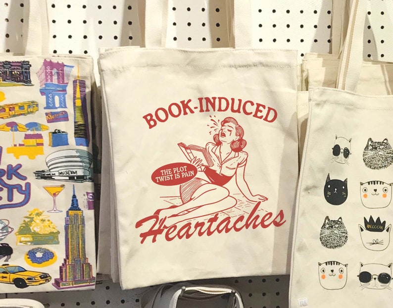 May include: A white canvas tote bag with a red graphic of a woman reading a book and the text "BOOK-INDUCED HEARTACHES" and "THE PLOT TWIST IS PAIN".