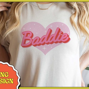 May include: A white t-shirt with a pink heart design that says "Baddie" in a retro font.