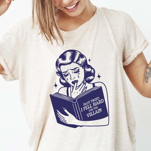 May include: A white t-shirt featuring a blue illustration of a woman reading a book. The book has the text "Plot Twist: I Fell Hard For The Villain".