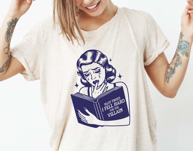 May include: A white t-shirt featuring a blue illustration of a woman reading a book. The book has the text "Plot Twist: I Fell Hard For The Villain".