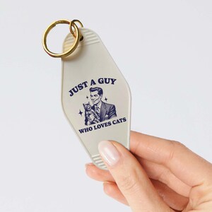 May include: White motel key chain with a gold key ring. The keychain has a retro-style illustration of a man holding a cat and the text "Just a guy who loves cats".