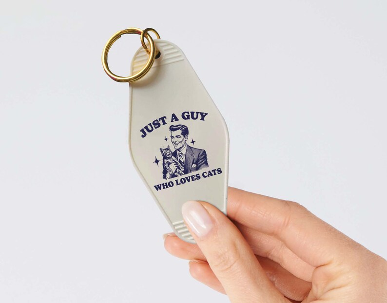 May include: White motel key chain with a gold key ring. The keychain has a retro-style illustration of a man holding a cat and the text "Just a guy who loves cats".