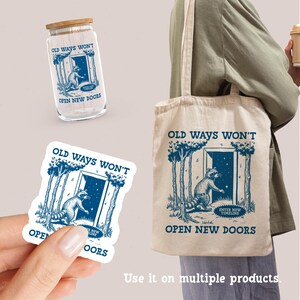 May include: A tote bag with a white background and blue text that reads "Old ways won't open new doors". The bag features a raccoon standing in front of a doorway with a starry sky behind it. The raccoon is holding a sign that reads "Enter new timeline".