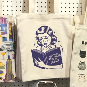 May include: A white tote bag featuring a black and white illustration of a woman reading a book. The book has the text "Plot Twist: I Fell Hard For The Villain".