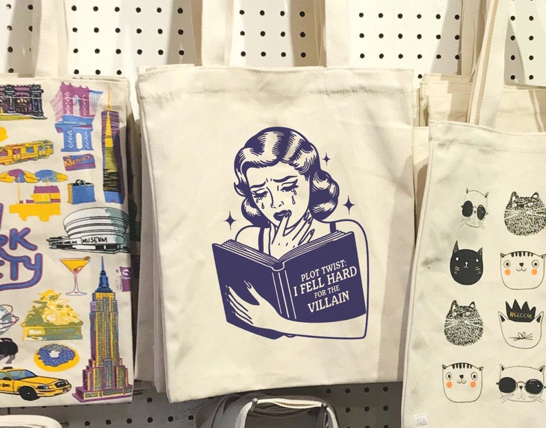 May include: A white tote bag featuring a black and white illustration of a woman reading a book. The book has the text "Plot Twist: I Fell Hard For The Villain".